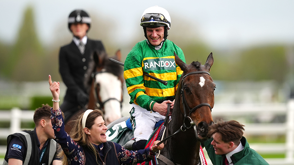 I am Maximus after his 2024 Grand National win, with jockey Paul Townend in green and gold on the horse and and three others around it on the ground