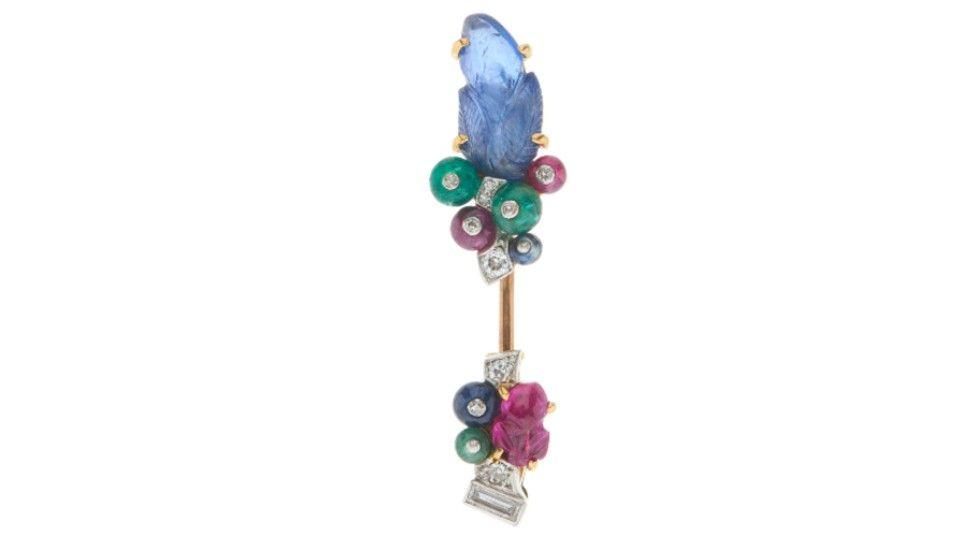 A brooch which contains two clusters of jewels on a piece of gold. At the top end, there is a cluster containing a large light blue gemstone, along with smaller diamonds and colourful gemstones, and at the bottom of the brooch, there is another cluster of gems in a similar arrangement, but with a smaller pink gemstone rather than a large blue one.