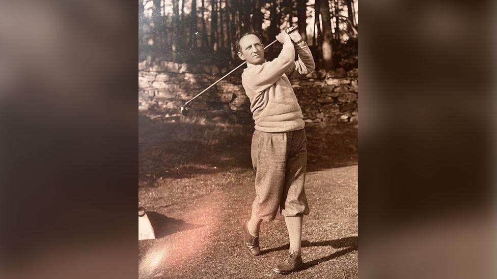 Old sepia image of golfer, Bobby Cruickshank, swinging golf club, with trousers tucked into socks, with a wall and trees in the background.