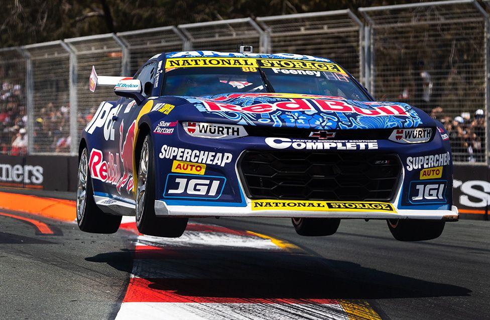 A blue and red race car with prominent Red Bull branding is airborne over a curb on a racetrack. The car features sponsor logos and metal fencing lines the track, spectators are visible in the background under sunny conditions.