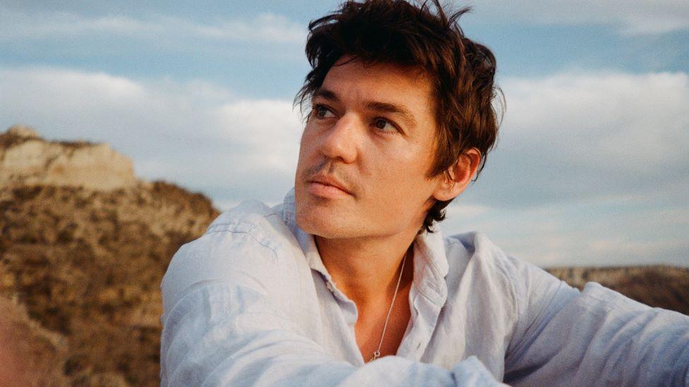 A man with brown hair in a white shirt sits outside by a grassy cliff and gazes into the distance.