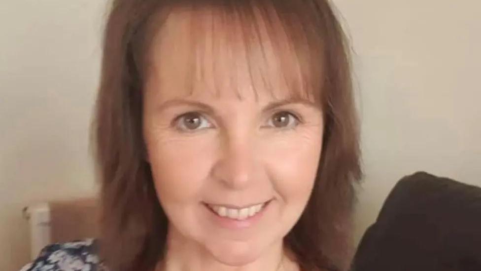 Heather Smedley, who has brown shoulder length hair, smiles while sat on a sofa.