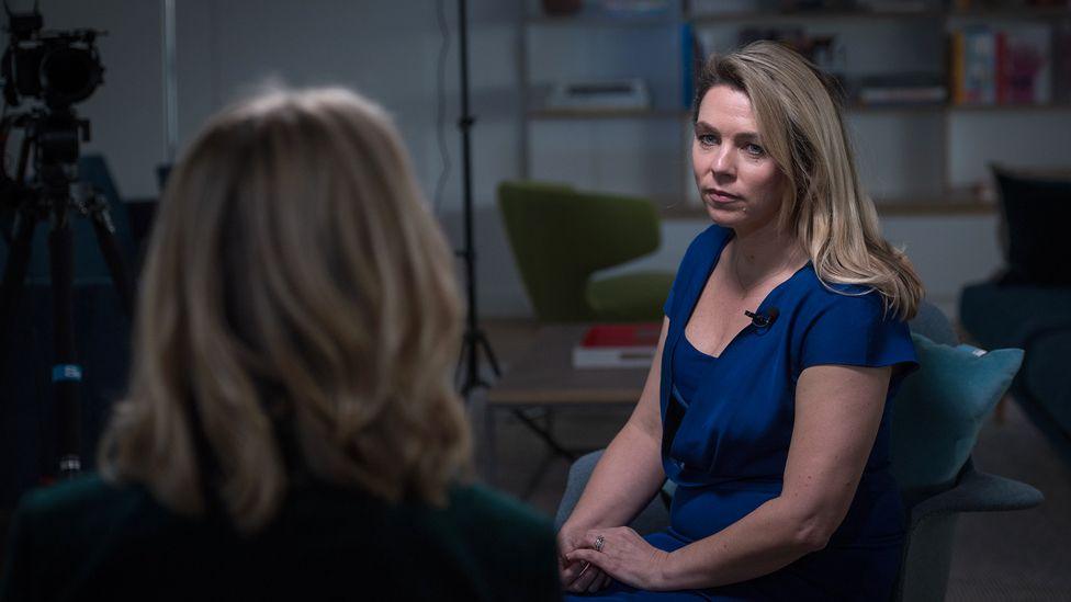 Sarah Wynn-Williams - a woman with long blond hair, wearing a blue dress - sits on a chair opposite the BBC's Katie Razzall, who has her back to the camera.