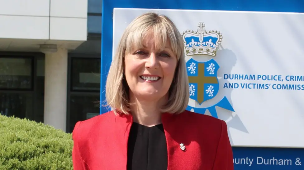 Joy Allen standing in front of a blue and white Durham Police sign. She has blonde short hair with a fringe and is wearing a red blazer with black top.