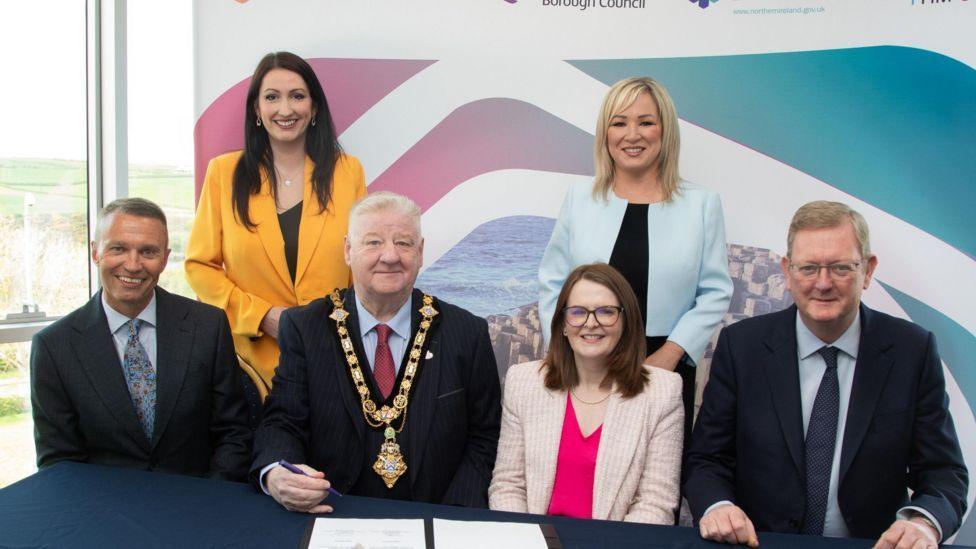 A number of officials including Northern Ireland's finance minister, and first and deputy first ministers sit at a table after singing a deal for the regeneration of causeway coast and glens council area.