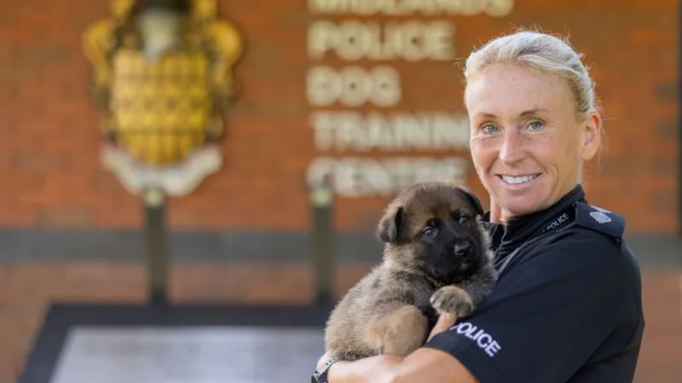 Insp Rachel Crump is wearing a black police uniform and holding a puppy. She is standing in front of the dog training centre.