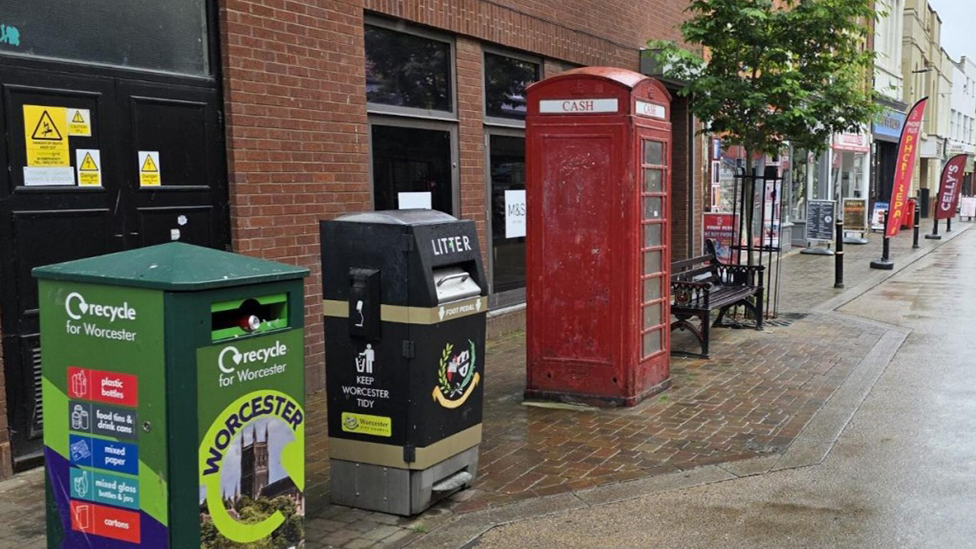 A red phone box on a street in Worcester. There is a black metal bench on the right of it and recycling and litter bins on the left. There are shops, signs and a small tree in the background