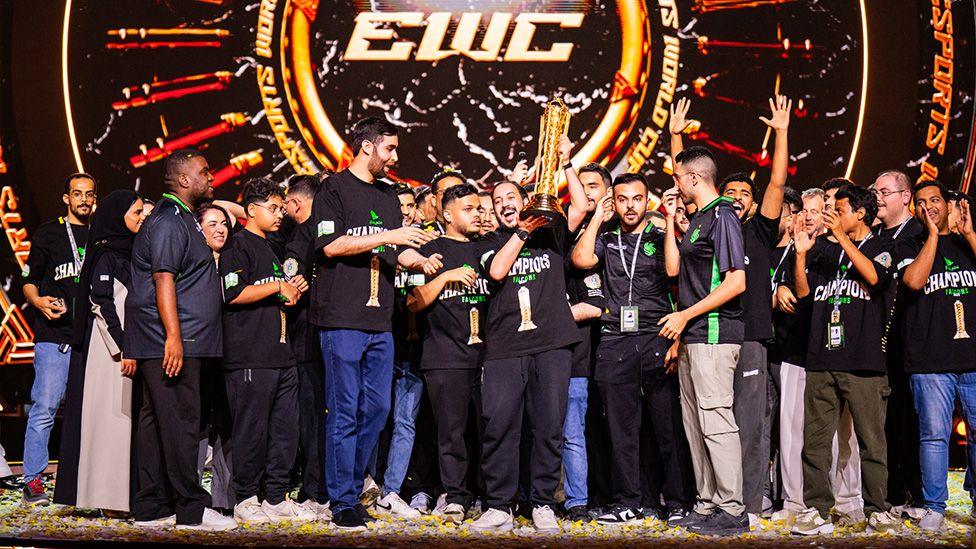 A large group of young men wearing black t-shirts with "Champions" printed on them stand on a stage littered with pieces of confetti as one member at the front of the group triumphantly holds up a tall golden trophy amid the scenes of celebration. Behind them an "EWC: Esports World Cup" logo is visible. 