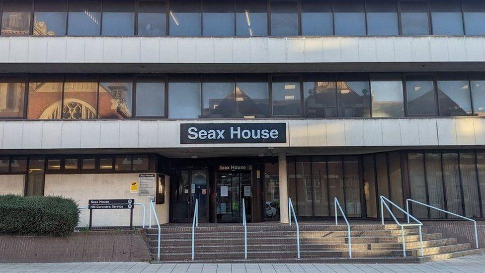 The entrance to Essex Coroner's Court at Seax House - a photo taken from the other side of the road. There are steps and white-painted railings leading up to the court entrance, and the sign on the 1960s multi-storey building reads SEAX HOUSE.