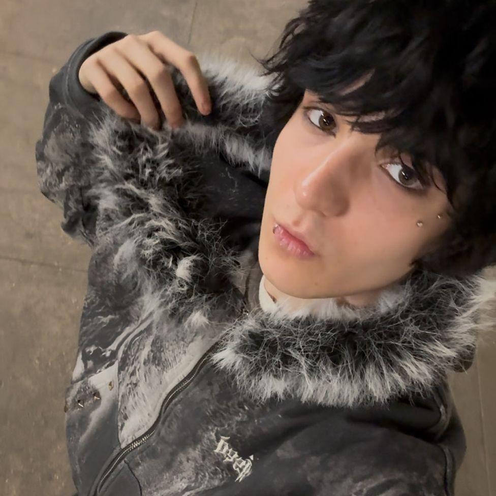 Ifesinachi Mamah, taking a selfie wearing a grey coat with a fluffy trim hood. They have black hair and a lip and face piercings.