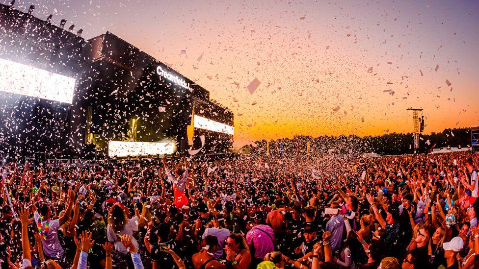 An artist performs on the main stage of Creamfields, which is bordered by huge white lights and stands in front of a huge crowd of people, who are looking skywards at a spray of confetti and the sunset