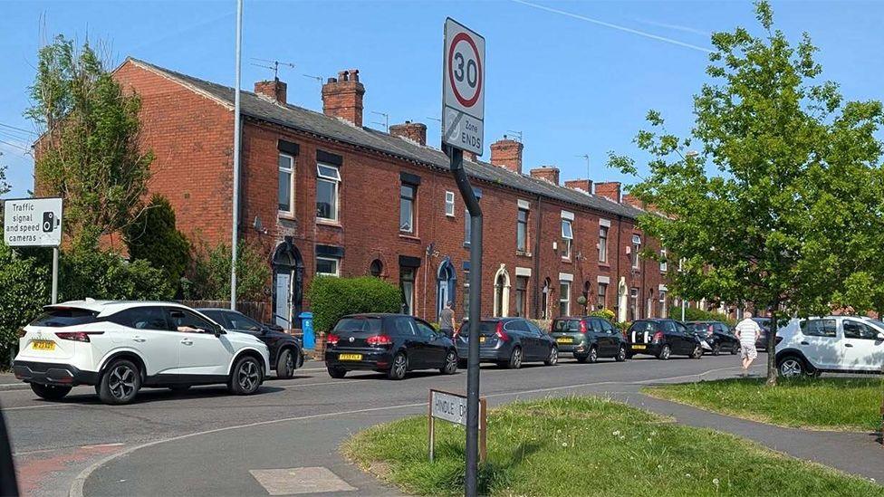 A queue of cars on a sunny day on a road in Royton going past a row of red brick terraced houses