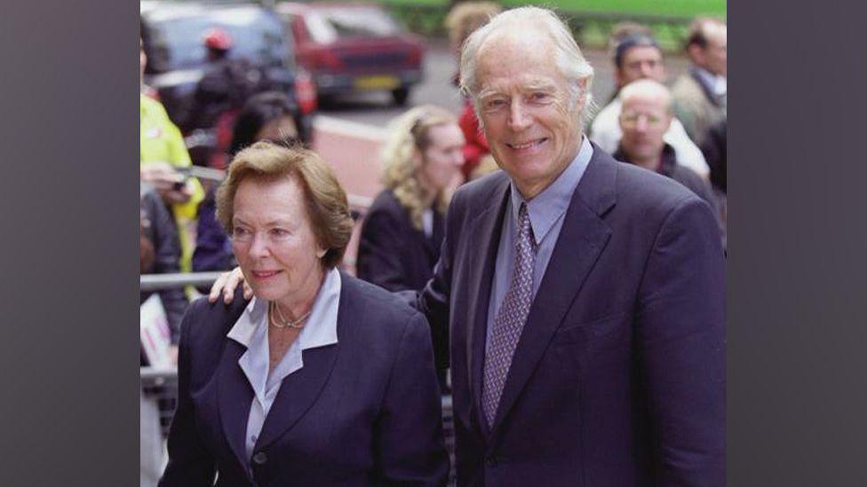 Sir George Martin and Lady Judy Martin are pictured together in 2000. Sir George is wearing a navy blue suit and light blue shirt with a purple spotted tie while his wife is wearing a navy suit jacket and light blue blouse.