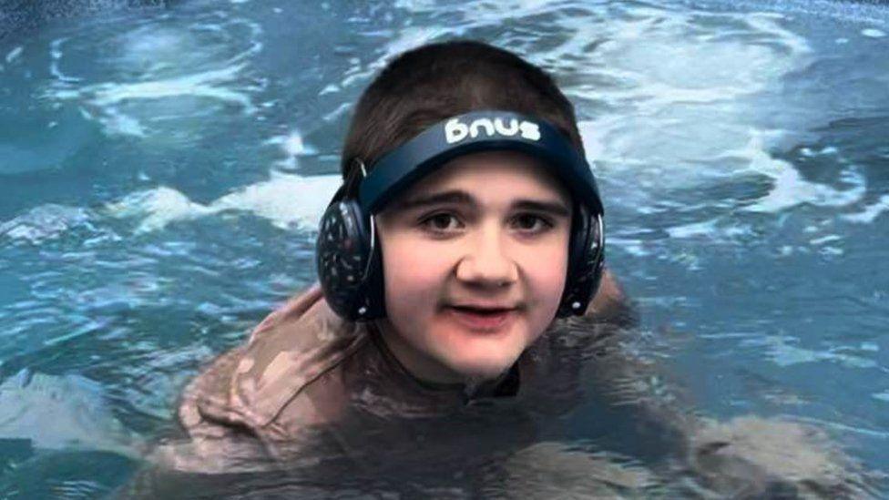 Tobie, in a swimming pool facing the camera. He is wearing a brown t-shirt and black ear defenders. 