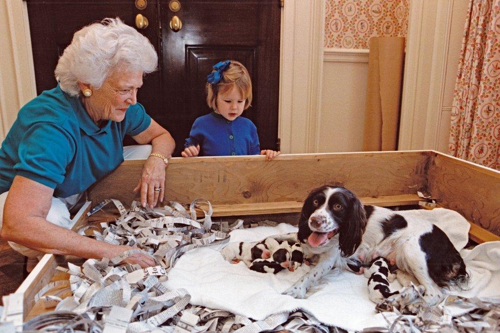 Barbara Bush and her granddaughter look at a dog who is feeding a littler of puppies