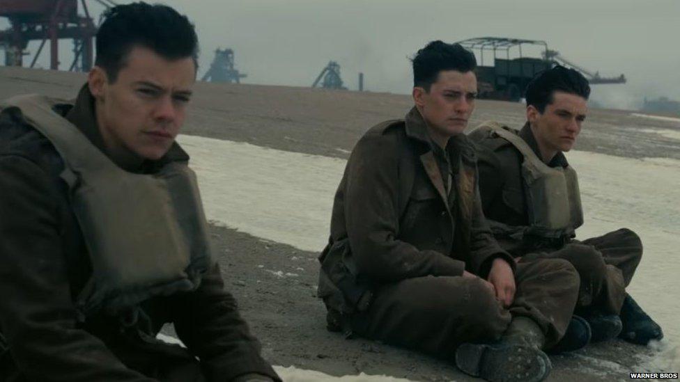 Harry Styles in Dunkirk Movie. He is sitting with co-stars on the beach.