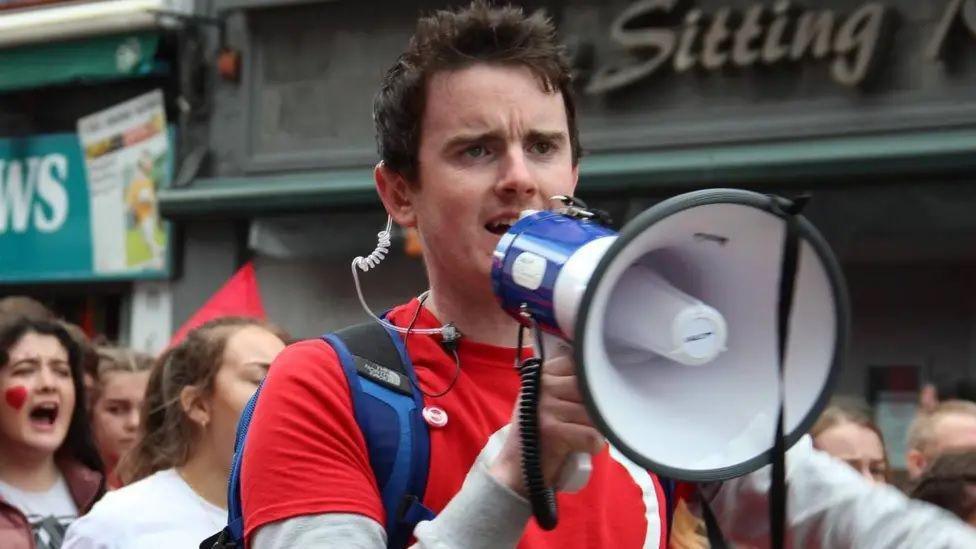 Pádraig Ó Tiarnaigh has short brown hair and is wearing a red t-shirt and blue backpack. He is speaking into a microphone and has a clear/white earpiece.