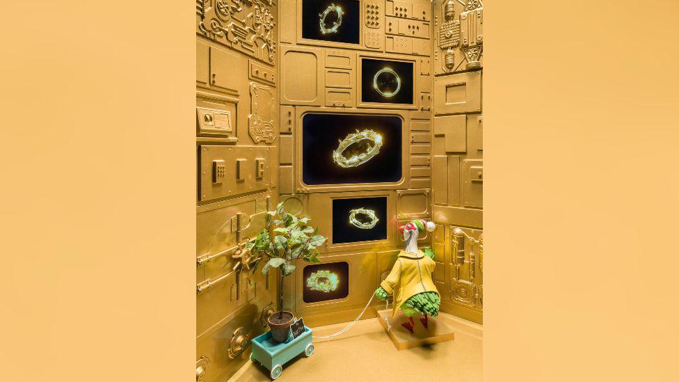 A scene with many animated objects. A yellow partridge is in a golden room looking at images of rings. There is a plant pot in the corner.