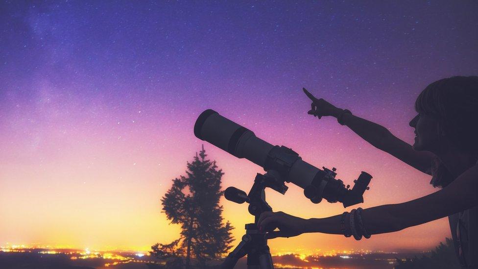 A person points at the sky while holding a telescope.