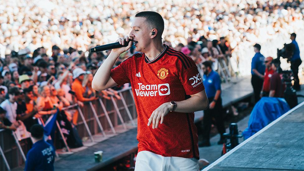 Aitch wearing the new Manchester United shirt, which is red and has the team logo and Adidas sponsor, along with the name of their Team Viewer sponsor in the middle of the shirt. He is wearing white trousers, and is holding a microphone, and has a watch on his left hand. The background has thousands of people watching Aitch perform.