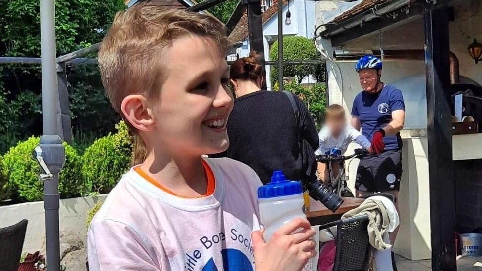 A head an shoulder shot of Max, pictured from his right side. He had short brown hair and is wearing a white T-shirt over and orange T-shirt. He is holding a clear water bottle with a blue lid and is smiling, broadly