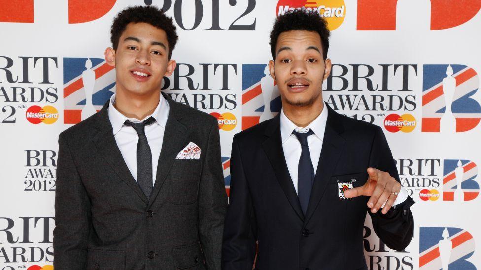 Jordan Stephens and Harley Alexander-Sule wearing suits and smiling at the camera. Behind them is signage for the 2012 Brit Awards. Both men are wearing black suits. 