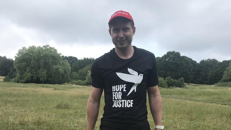 Aaron Robinson, aged 40, smiles at the camera wearing a black t-shirt with the Hope for Justice's charity branding. Behind him is a grassy field where he runs.