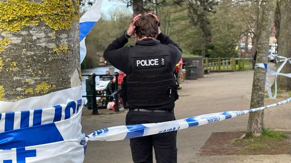 Police officer walking towards river with blue and white police emergency tape behind him, Norwich