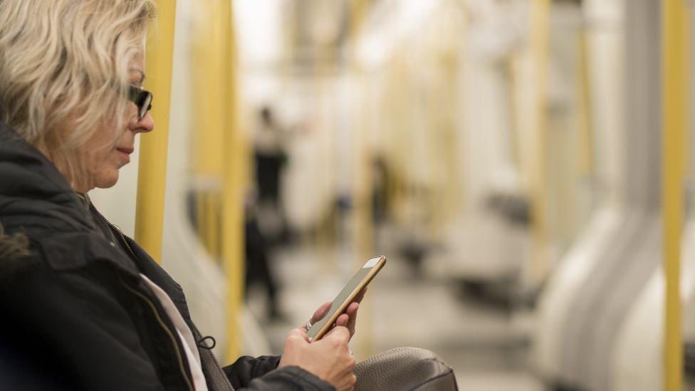 A woman with blonde hair in a black coat sits on the Tube and looks at her phone. The carriage is largely empty.