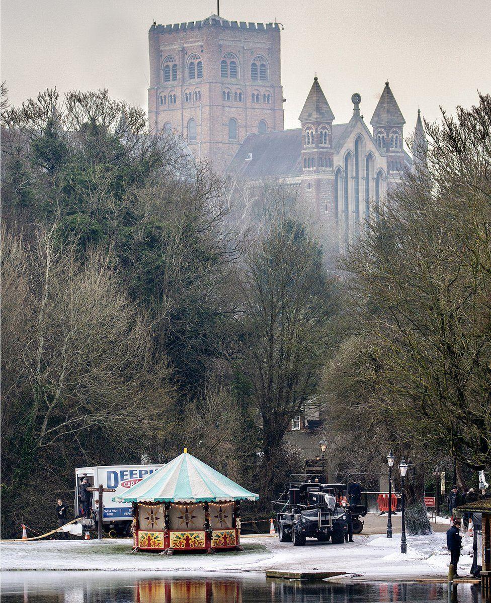 A fairground stall can be seen next to St Albans Verulamium lake with fake snow on the ground with trees and St Albans Cathedral in the backgorund.