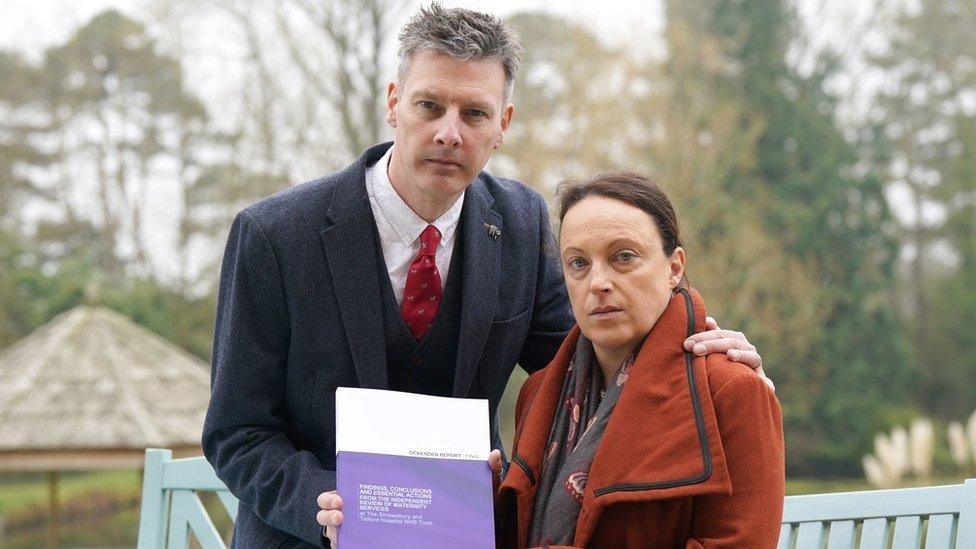 A man in a dark suit with greying spiky hair, and a woman with tied-back brown hair and wearing an orange-brown coat hold a purple and white document titled "Ockenden Report".
