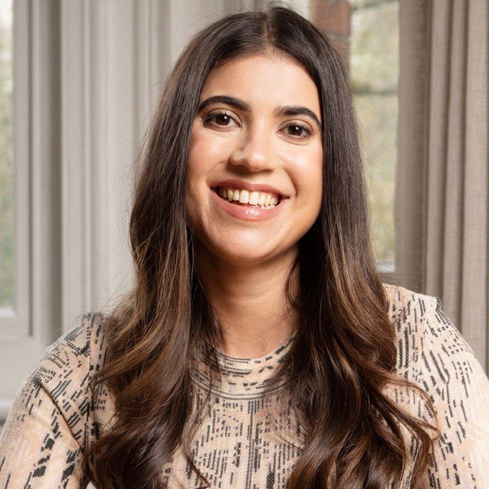 A woman in a patterned brown and black top with long brown, curly hair sits in front of a window