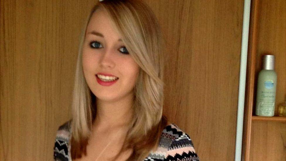 Close up of Katie Allan - a young blonde woman smiling in front of a wood panelled wall.