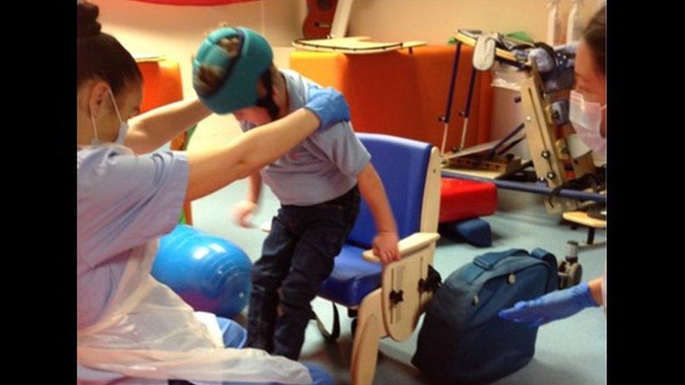 A pupil practising his sit-to-stand routine with his new chair with a teacher in PPE mask and gown assisting him.