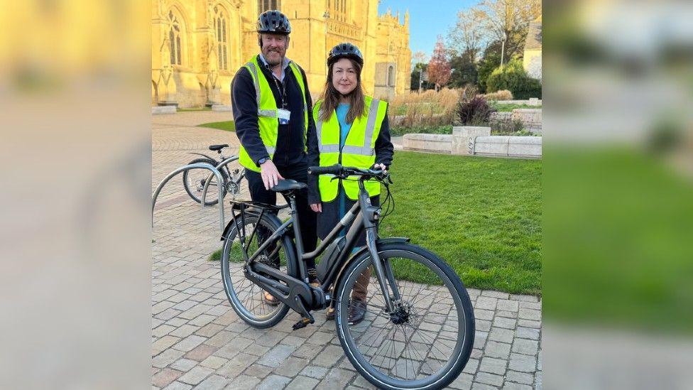 Councillors David Gray and Chloe Turner standing in front of Gloucester cathedral with an e-bike and wearing bicycle helmets and hi-vis jackets.