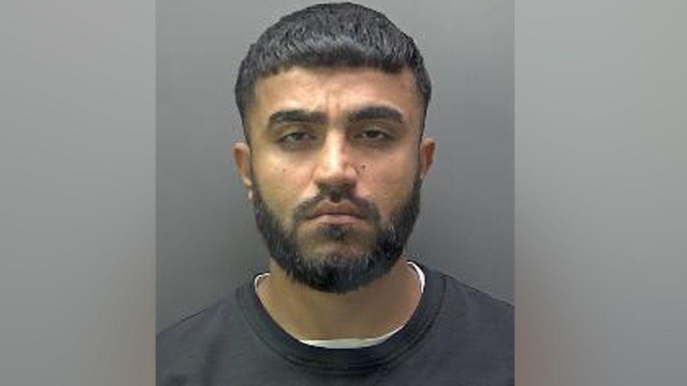 A mugshot of Galmne, wearing a white t-shirt under a black sweater. He has a black beard and moustache and short dark hair.