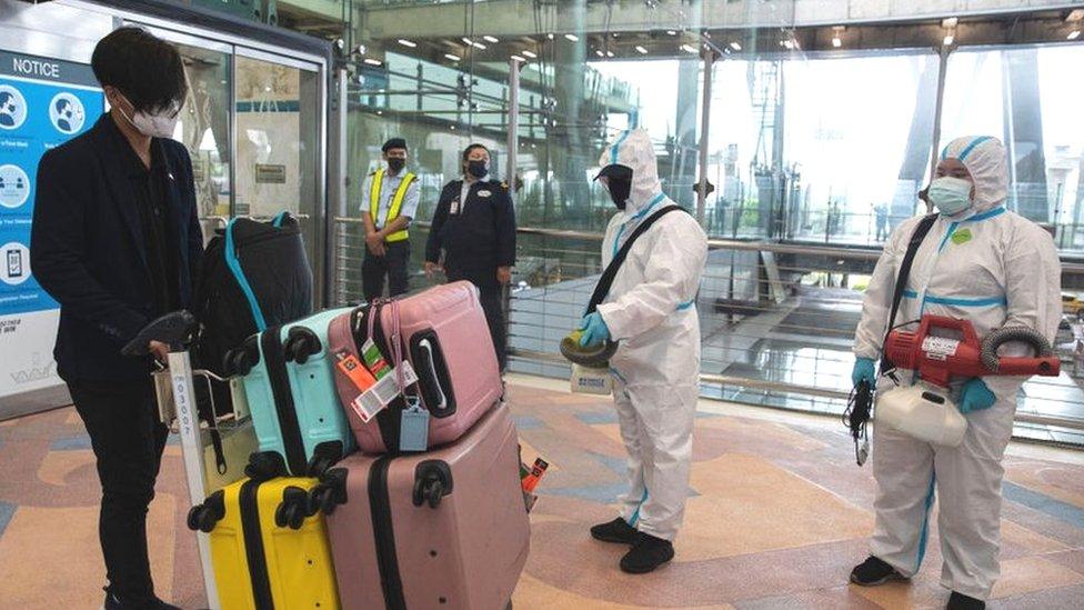 Suvarnabhumi airport staff wearing Personal protective equipment (PPE) suits spray sanitizers on a passenger's luggage on arrival at the airport.