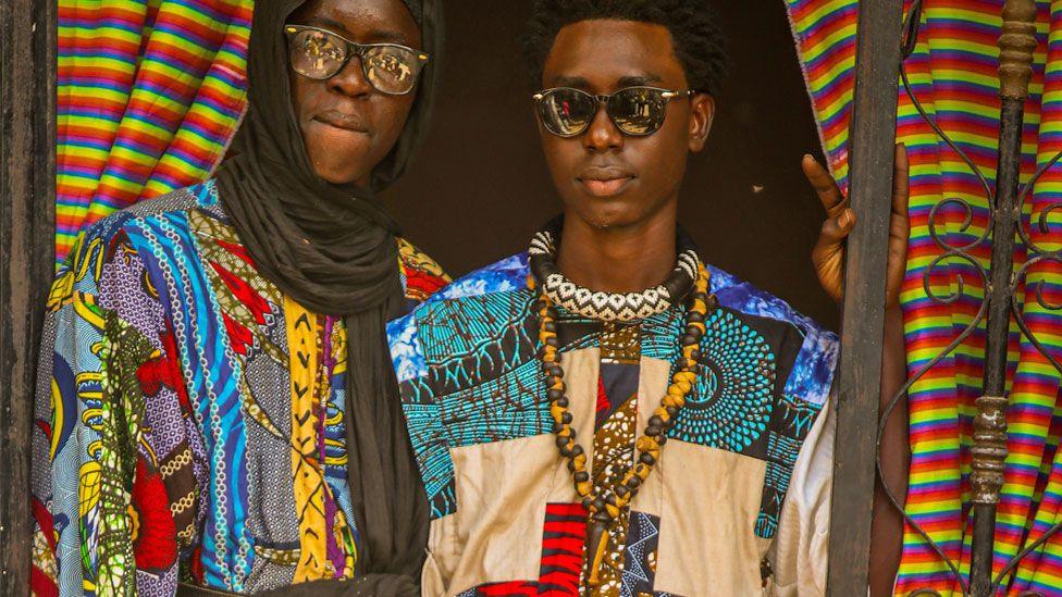 Two men in multi-patterned colourful patchwork clothes look at the camera framed by a doorway. One is wearing a black headscarf and glasses, the other is in sunglasses and has beaded necklaces around his neck - in Diourbel, Senegal, April 2024