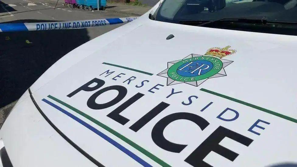 The white car bonnet of a Merseyside Police patrol car, bearing the name of the force in black and blue lettering. Police tape can be seen across a road behind the vehicle.