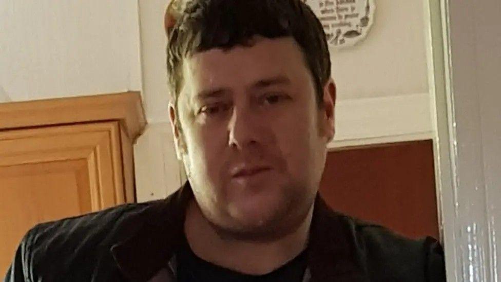 Ian Staves standing in a door way looking towards a camera. He has brown hair and is wearing a dark jacket.