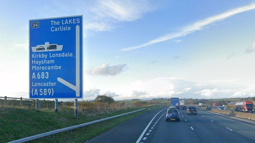 Street view image of the M6 motorway junction just before junction 34, showing a blue motorway sign to the left pointing north to the Lakes and left to the Heysham ferry