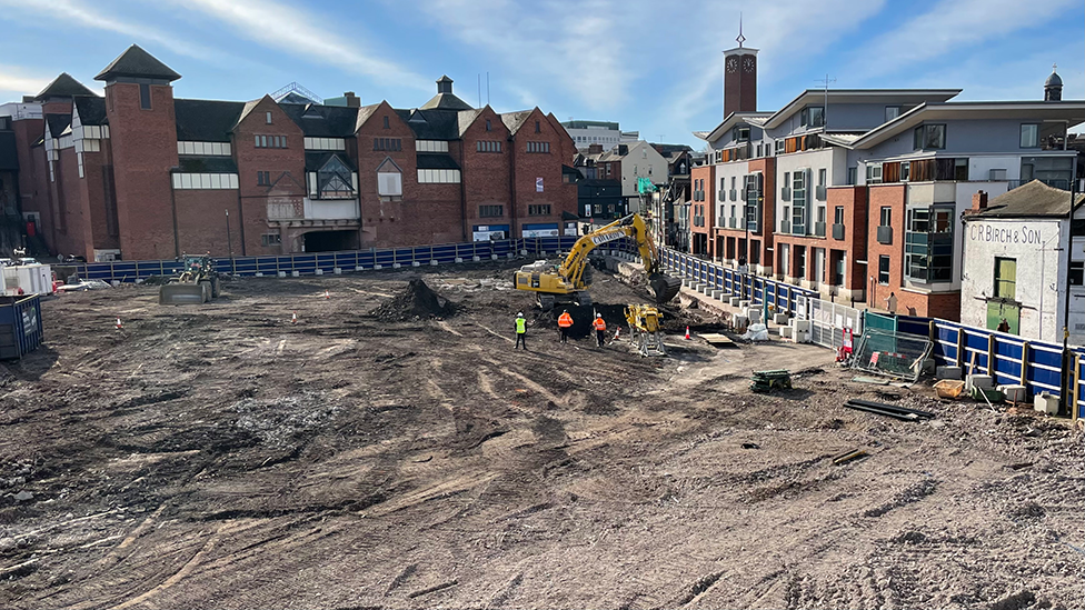A large, open building site, viewed from a raised position, bordered on two sides by tall buildings, in Shrewsbury town centre. There are people wearing high visibility clothing stood by a yellow digger. There is fencing around the site, with a gate in the right hand side of the site.