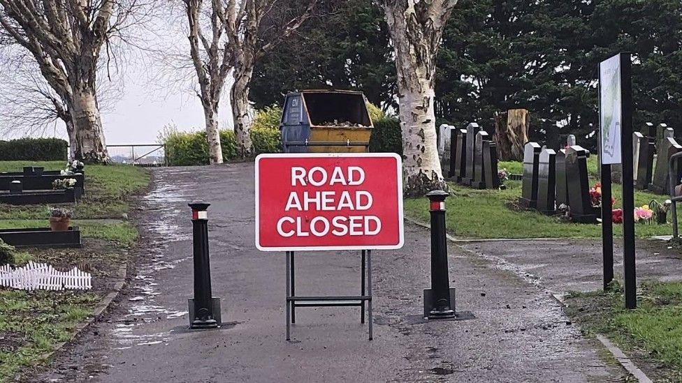 Two metal bollards and a red road sign which reads "road ahead closed"  are blocking a road through a cemetery. Headstones are visible either side and a skip can be seen in the background.  