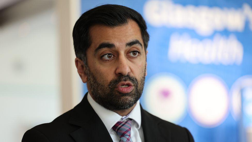 A man with a dark hair and beard speaks in front of a blue sign. He is wearing a black suit, white shirt and purple tartan tie. 