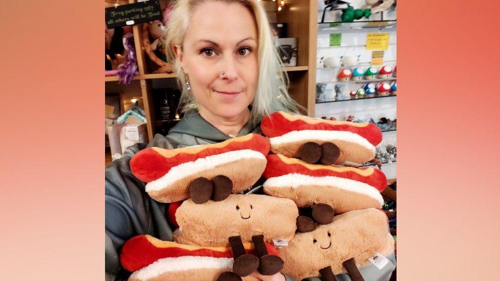 A blonde haired woman with a nose stud and her hair tied up holding several hot dog toys with smiley faces - they are Jellycat toys. You can see shelves full of items in the background.