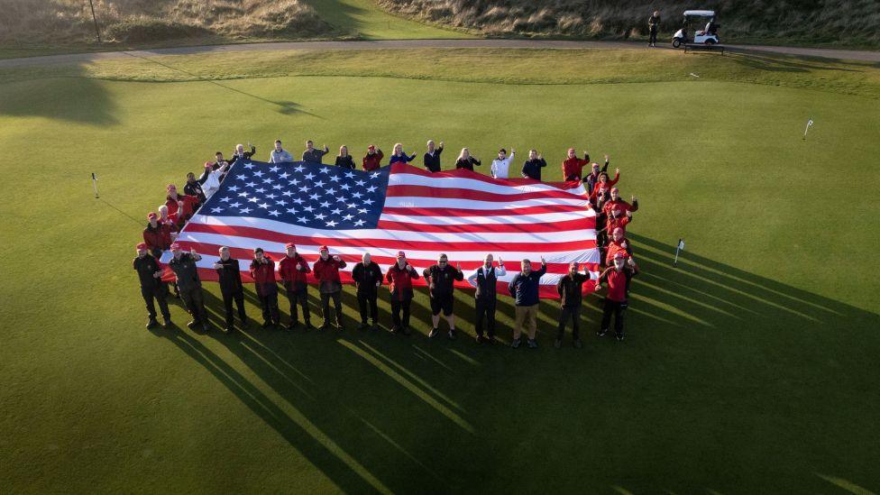 A large US flag with stars and stripes held up by dozens of golf course staff standing on a green, looking up and waving to a camera