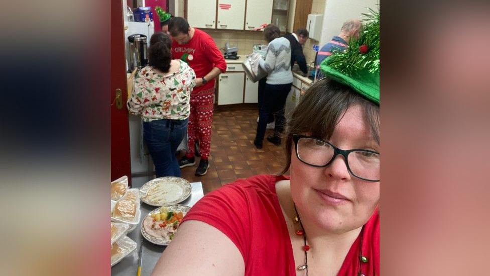 Rebecca is taking a selfie - she has brown hair tied up and black glasses. She is wearing a re t-shirt and a green christmas tree hat. In the background is a kitchen with people working, while dressed in christmas themed outfits. There is food on plates covered in cling film on a table to Rebecca's left