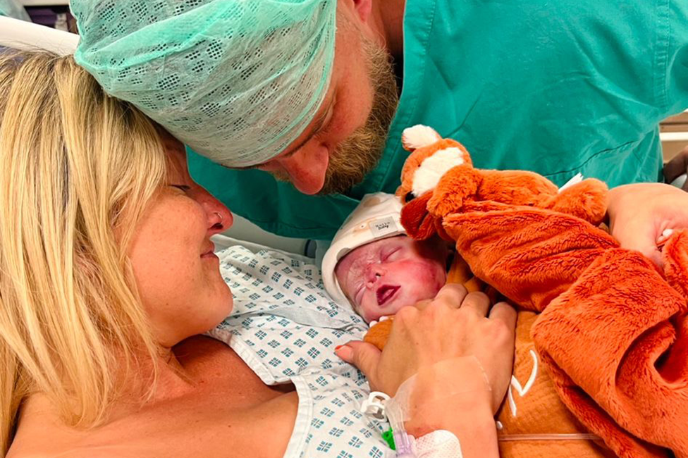 Andrea McCartney in a hospital gown holding her newborn baby Kit, with a smile on her face. Her partner is in green surgical scrubs close to his son