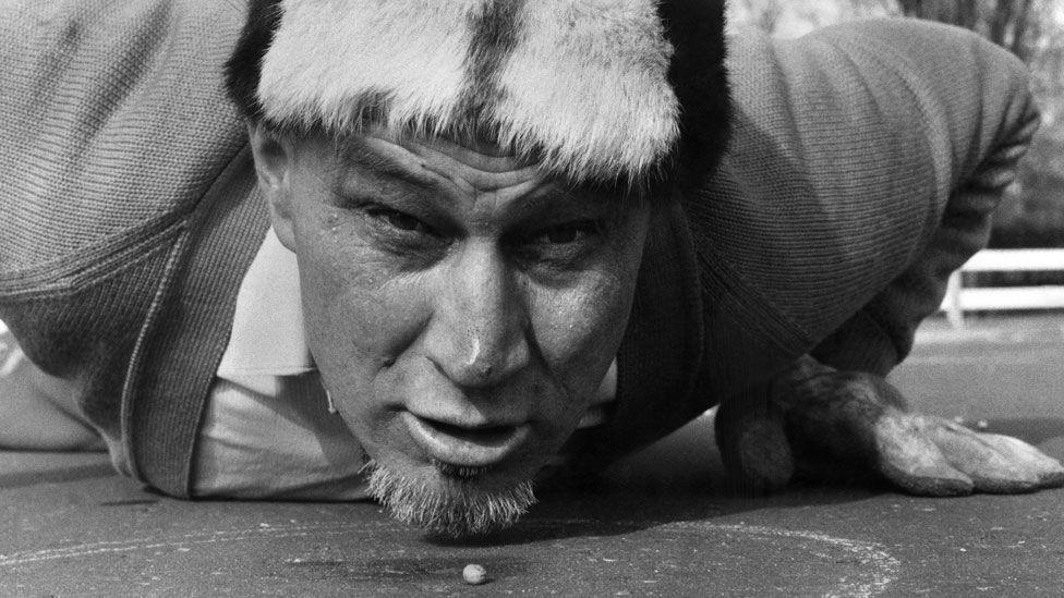 A black and white image of Walter Cornelius face on and flat on his stomach with his hands beneath his shoulders. His head is raised and he is looking forward and in front of him on the ground is a pea.