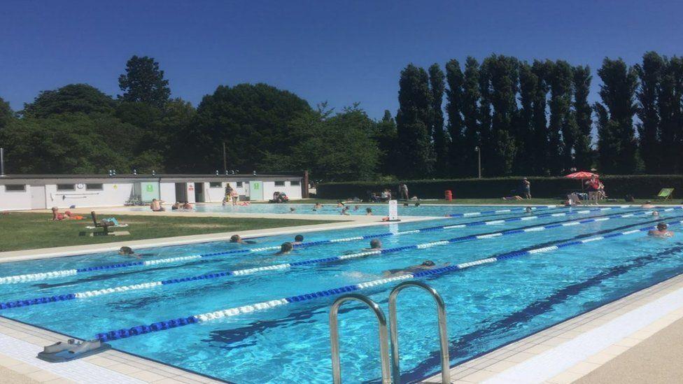 Open-air pool with blue and white lane ropes set, lane swimming boards in place and lifeguard highseat to the side set in park